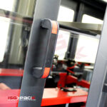 Zbliżenie czarno-czerwonej klamki na przezroczystych drzwiach, w tle Automatyczna formierka Speed Former 50 SB Servo z czerwonymi i czarnymi elementami. Logo ISG PACK jest widoczne w lewym dolnym rogu.