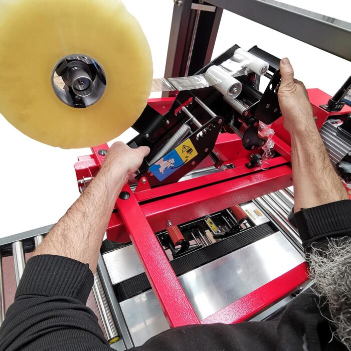 Osoba ładuje dużą rolkę przezroczystej taśmy na półautomatyczną zaklejarkę Easy Tape 50 TBB, regulując mechanizm ręcznie.