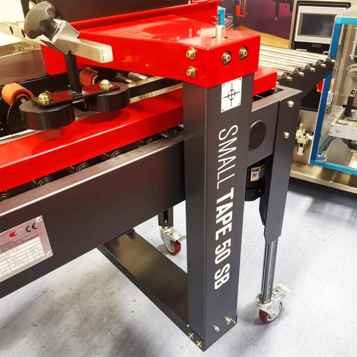 Zbliżenie na Półautomatyczną zaklejarkę Small Tape 50 SB z białą etykietą na czarnej metalowej ramie z czerwonymi i czarnymi elementami, umieszczoną na kółkach na szarej podłodze.