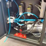 Zbliżenie przemysłowego panelu sterowania wewnątrz metalowej obudowy z oznaczonymi zaciskami, niebieskimi i czarnymi przewodami oraz metalowym cylindrem na czerwonym wsporniku - część systemu Automatyczna owijarka palet Ultra Wrap 2000 BWF.