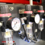 Zbliżenie pneumatycznego manometru i regulatora z przezroczystymi rurkami na czarnym panelu. W lewym dolnym rogu widoczne jest logo Automatyczna zaklejarka Matic Tape 50 CF HM.