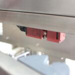 Zbliżenie czerwonego czujnika bezpieczeństwa zamontowanego na Automatycznej zaklejarce Auto Tape 50 CF SS, z widocznymi śrubami i etykietami, używanego do automatyzacji i bezpieczeństwa.