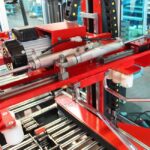 Zbliżenie Automatycznej zaklejarki Matic Tape 50 CF w fabryce, pokazujące jej czerwoną metalową ramę, wiele metalowych prętów, silnik, cylindry hydrauliczne lub pneumatyczne, widoczne przewody i rurki.