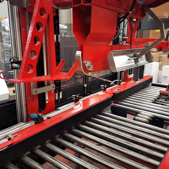 Zbliżenie automatycznej zaklejarki Matic Tape 50 CF w kolorze czerwonym, umieszczonej na metalowym przenośniku rolkowym w fabryce, podkreślające jej części mechaniczne i rolki.