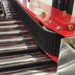 Zbliżenie na czarny system przenośnika taśmowego z metalowymi rolkami i czerwonymi elementami, prezentujący Półautomatyczną zaklejarkę Easy Paper 50 SB, gdy taśma gładko przesuwa się po rolkach.