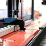 Zbliżenie czujnika mechanicznego i metalowego ramienia na Automatycznej formierce Speed Former 50 SB Servo czerwony sprzęt przemysłowy; logo ISG PACK pojawia się w lewym dolnym rogu.