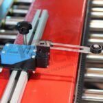 Zbliżenie niebieskiego wyłącznika krańcowego z dźwignią rolkową, zamontowanego na metalowej szynie przymocowanej do czerwonej powierzchni w pobliżu rolek przenośnika w Automatycznej zaklejarce Matic Tape 50 CF HM.