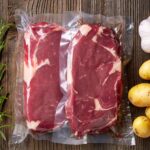 Dwa surowe steki, uszczelnione próżniowo za pomocą Przemysłowej pakowarki próżniowej Easy Vac 500D FT, leżą na drewnianej powierzchni w otoczeniu świeżego rozmarynu, czosnku i żółtych ziemniaków.