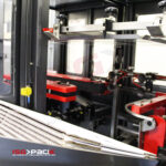 Zbliżenie na maszynę pakującą z automatyczną formierką Speed Former 50 SB XL z metalowymi ramionami i czerwonymi komponentami, ułożoną tekturą falistą oraz logo Isopack z napisem "automatyczny system pakowania" w lewym dolnym rogu.