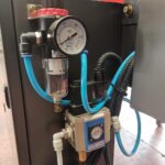 Zbliżenie regulatora ciśnienia powietrza z manometrem (w MPa), niebieską rurką, filtrem i okablowaniem elektrycznym zamontowanym na metalowym panelu Automatycznej zaklejarki Auto Tape 50 HM.