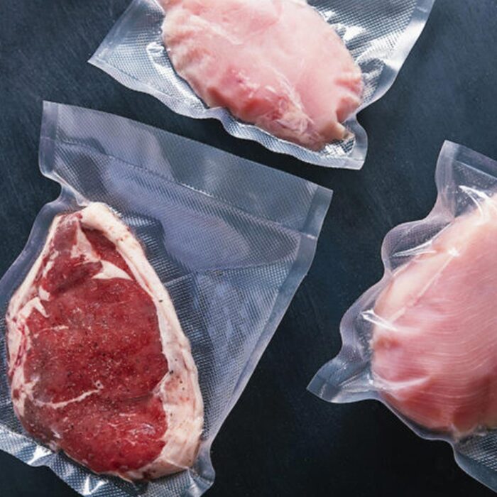 Trzy kawałki surowego mięsa, zarówno czerwonego, jak i białego, są zamykane próżniowo w przezroczystych plastikowych torebkach za pomocą Przemysłowej pakowarki próżniowej Easy Vac 420S FT i eksponowane na ciemnej powierzchni.