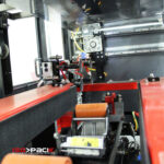 Zbliżenie automatycznej maszyny pakującej Speed Former 50 SB XL z czerwono-czarnymi częściami, przenośnikami i rolkami. W lewym dolnym rogu znajduje się logo IsoPack i napis "Automatic Packaging System".