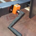 Zbliżenie czarnej metalowej korby na pomarańczowym elemencie mechanicznym Automatycznej Formierki Speed Former 50 SB Servo, z betonową podłogą w tle.