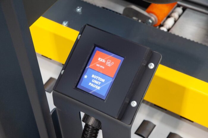 Zbliżenie panelu sterowania Nastro-SB AQUA półautomatyczna zaklejarka kartonów z kolorowym ekranem dotykowym wyświetlającym kod błędu E23 i opcję BOTTOM UNIT PAUSE; w tle widać przenośnik taśmowy i części mechaniczne.