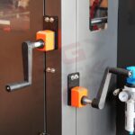 Dwie czarne korby ręczne z pomarańczowymi podstawami są zamontowane z boku Automatycznej Formierki Speed Former 50 SB XL, w pobliżu szklanego okienka i metalowej rurki z niebieskim zaworem.