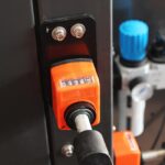 Zbliżenie pomarańczowego licznika mechanicznego z odczytem 00036, przykręconego do metalowej powierzchni na Automatycznej Formierce Speed Former 50 SB XL, z pokrętłami sterującymi i niebieskim zaworem widocznymi w tle.