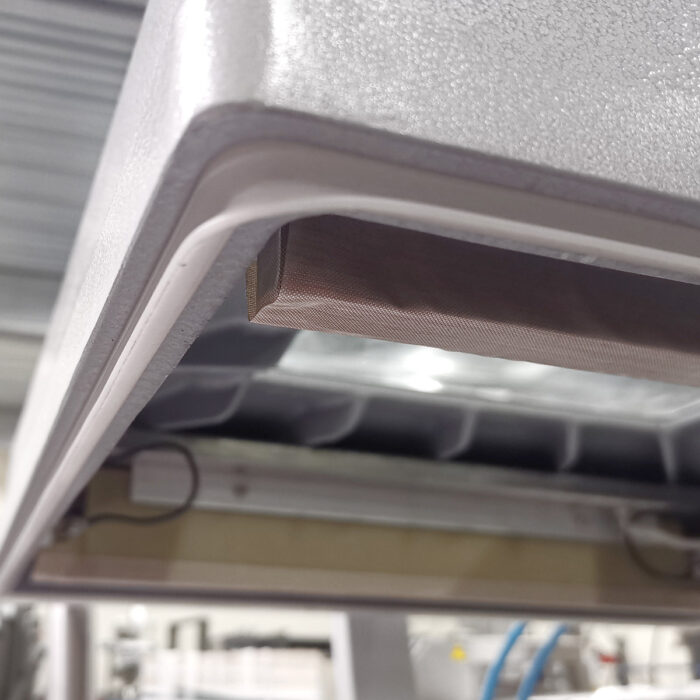 Zbliżenie metalowej części maszyny z brązowym prostokątnym paskiem pod spodem, prawdopodobnie z Komorowej pakowarki próżniowej Ultra Vac 550 SP, pokazanej w otoczeniu przemysłowym z rozmytymi maszynami w tle.