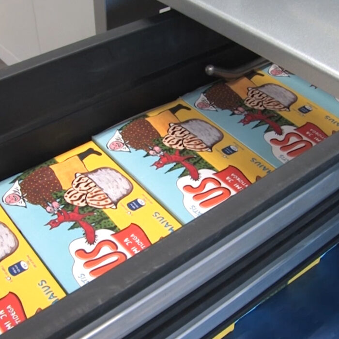 Zbliżenie na wiele zapakowanych bochenków chleba w metalowej szufladzie, zaplombowanych Automatyczną zgrzewarką do zgrzewek FA800AR 280ST, z kolorowymi etykietami z ilustracjami chleba i owoców.