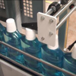 Butelki wypełnione niebieskim płynem i białymi nakrętkami przesuwają się wzdłuż przenośnika taśmowego w zakładzie pakowania przemysłowego, gdzie Automatyczna zgrzewarka do zgrzewek FA800AR-150Z zapewnia bezpieczne owijanie.