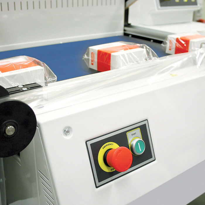Zbliżenie na półautomatyczną zgrzewarkę kątową Flexy Sealer 55A z pudełkami owiniętymi folią na przenośniku taśmowym; jej panel sterowania zawiera duży czerwony przycisk zatrzymania awaryjnego i zielony przycisk startu.