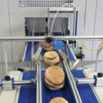 Stosy chleba pita przesuwają się wzdłuż niebieskiego przenośnika taśmowego przez Automat pakujący poziomy FLOWPACK SBM550S-LR w zakładzie przetwórstwa spożywczego, z widocznymi metalowymi prowadnicami i czujnikami.