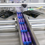 Fioletowe cylindryczne elementy są ustawione w rzędzie, poruszając się wzdłuż metalowego przenośnika z automatyczną pakowarką flow-pack SR550 w zakładzie produkcyjnym lub pakującym.