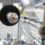 Zbliżenie na Automat pakujący poziomy FLOWPACK SBM750S pokazujące metalowe rolki, czarną szpulę i różne elementy mechaniczne używane w zautomatyzowanej linii produkcyjnej.