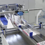 Zbliżenie na Automat pakujący poziomy FLOWPACK SI550S-LR skutecznie sortujący i przenoszący zadrukowane arkusze z etykietą EXAMEN FERNÁNDEZ na przemysłowej linii produkcyjnej.