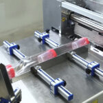 Zbliżenie na Automatyczną pakowarkę flow-pack SR750E owijającą cylindryczny przedmiot z czerwonymi końcami w przezroczystą folię z tworzywa sztucznego, prowadzoną przez regulowane niebieskie i srebrne szyny.