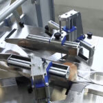 Zbliżenie Automatycznej pakowarki flow-pack SR550E pokazuje metalowe ramiona i rolki prowadzące i zgrzewające przezroczystą folię z tworzywa sztucznego wokół produktów na linii produkcyjnej.