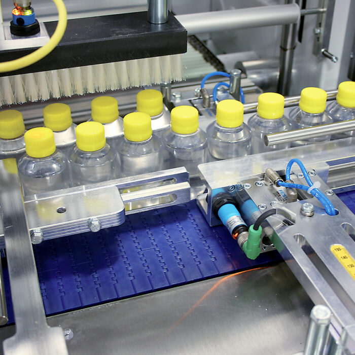 Plastikowe butelki z żółtymi nakrętkami przemieszczają się na przenośniku w zautomatyzowanej rozlewni, gdzie Automatyczna zgrzewarka do zgrzewek FA802AR 230R zgrzewa wielopaki wśród zaawansowanych maszyn i czujników.
