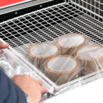 Używając Zgrzewarki kloszowej do folii Easy Sealer 85, osoba owija cztery rolki brązowej taśmy pakowej przezroczystą folią plastikową na powierzchni z siatki drucianej.