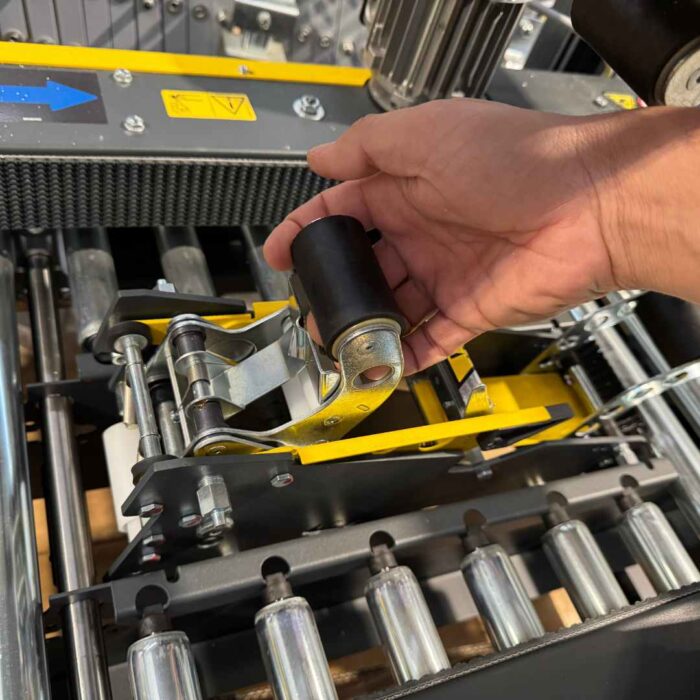 Ręka reguluje czarną rolkę na żółtym i metalowym Półautomatyczna zaklejarka kartonów SIAT S8, z widocznymi rolkami przenośnika i częściami mechanicznymi.