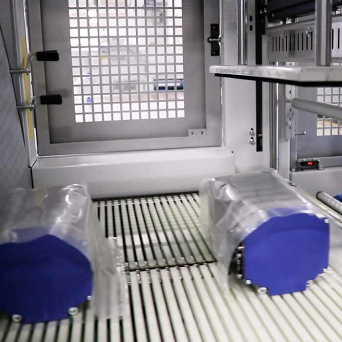 Dwa niebiesko-srebrne urządzenia elektroniczne owinięte w przezroczysty plastik znajdują się na przenośniku taśmowym wewnątrz Automatycznej zgrzewarki do pakietów i zgrzewek FA1102AS, z metalowymi drzwiami kratowymi widocznymi w tle.