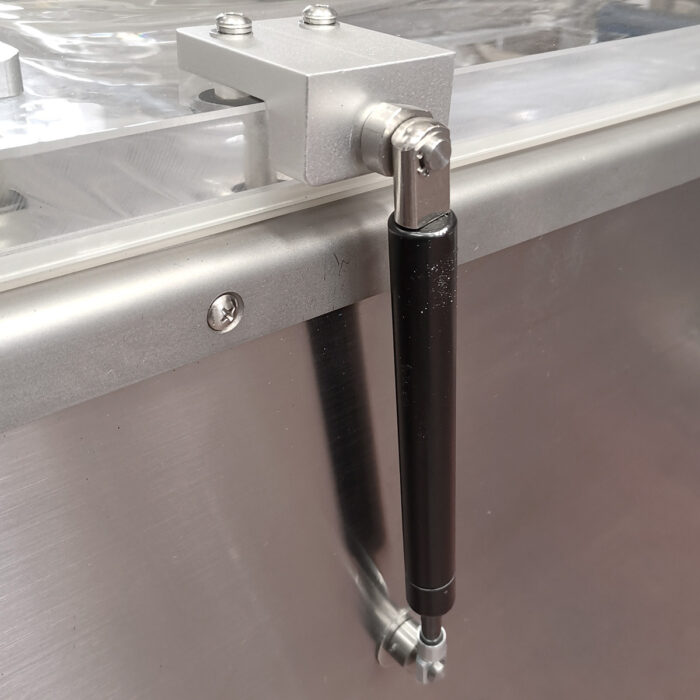 Zbliżenie metalowego zawiasu z czarną hydrauliczną kolumną gazową przymocowaną do powierzchni ze stali nierdzewnej przemysłowej pakowarki próżniowej Easy Vac 600S FT.
