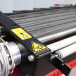 Zbliżenie na rolki przenośnika przemysłowego z żółtymi znakami ostrzegawczymi i strzałką kierunkową, część systemu pakowania Tunel obkurczający S 55 DC. W prawym górnym rogu znajduje się logo ISG PACK.