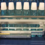 Sześć plastikowych butelek wypełnionych niebieskim płynem i zamkniętych białymi nakrętkami przesuwa się w rzędzie na przenośniku taśmowym w kierunku automatycznej zgrzewarki do zgrzewek FA800AR-150Z w sekcji maszyn zautomatyzowanych.