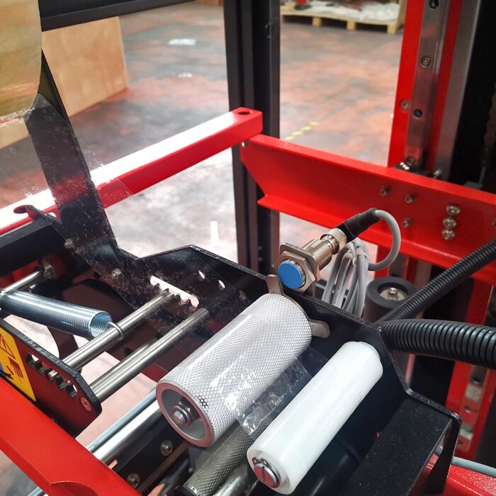 Zbliżenie na wewnętrzny mechanizm Automatycznej zaklejarki Matic Tape 50 CF z metalowymi rolkami, ostrzem tnącym i przewodowymi czujnikami na czerwonej ramie w magazynie przemysłowym.