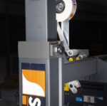 Zbliżenie na półautomatyczną zaklejarkę kartonów NASTRO-SB - R5-R6 nakładającą etykiety samoprzylepne; pomarańczowe, białe i czarne logo widoczne na ramie maszyny, w tle urządzenia przemysłowe.