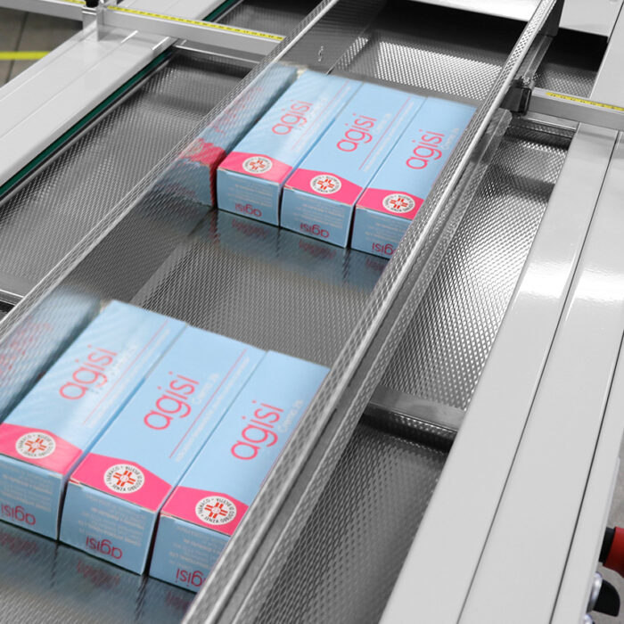 Kilka pudełek z etykietą agisi przesuwa się wzdłuż przenośnika taśmowego w zakładzie pakowania, gdzie Automatyczna zgrzewarka kątowa Ultra Sealer 70 HSS bezpiecznie zgrzewa niebieskie pudełka z różowymi i białymi akcentami.