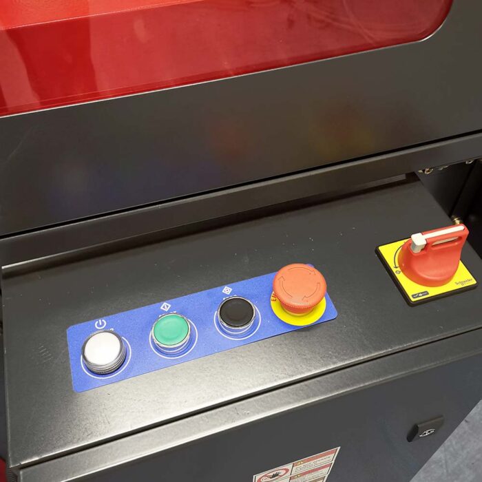 Zbliżenie na panel sterowania Automatycznej zaklejarki Auto Tape 50 CF ze srebrnym przyciskiem zasilania, zielonymi i czarnymi przyciskami, dużym czerwonym przyciskiem zatrzymania awaryjnego oraz czerwonym wyłącznikiem bezpieczeństwa z uchwytem na żółtej podstawie.