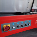 Zbliżenie na czerwoną Półautomatyczną bandownicę stołową Eco Strap 501L z otwartą pokrywą i panelem sterowania zawierającym pokrętła zasilania, timera, naprężenia, czasu chłodzenia i podawania/odwracania wraz z żółtymi i czerwonymi przełącznikami zasilania.