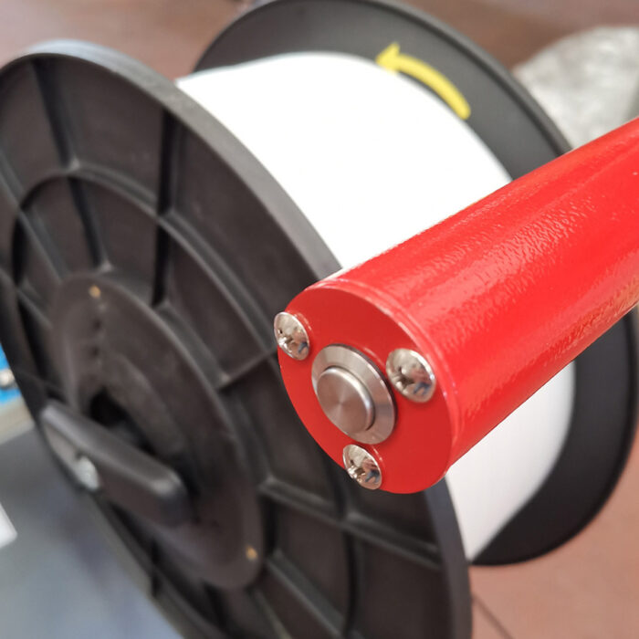 Zbliżenie Półautomatyczna bandownica do palet Pallet Strap T45: czarna szpula z białą taśmą, czerwony metalowy pręt zabezpieczony czterema śrubami i centralną śrubą oraz żółta strzałka na szpuli wskazująca kierunek obrotu.
