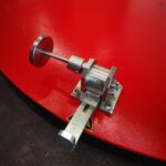 Zbliżenie na błyszczącą metalową dźwignię przykręconą do czerwonej maszyny przemysłowej, będącej częścią palety Półautomatyczna owijarka PRE-STRETCH Easy Wrap 2000 A RC, z częściowo widoczną naklejką ostrzegawczą pod spodem.
