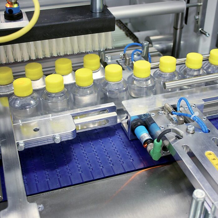 Przezroczyste plastikowe butelki z żółtymi nakrętkami przemieszczają się na przenośniku taśmowym przez zautomatyzowany zakład, mijając maszyny - w tym Półautomatyczną zgrzewarkę do zgrzewek FS800 - z metalowymi i niebieskimi częściami.