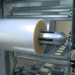 Zbliżenie dużej rolki przezroczystej folii plastikowej na metalowym wrzecionie wewnątrz maszyny przemysłowej, część procesu pakowania w Automat pakujący poziomy FLOWPACK SBM550S.