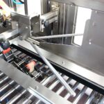 Zbliżenie na Automatyczną zaklejarkę Auto Tape 50 CF SS z konstrukcją ze stali nierdzewnej, wieloma prętami, dźwigniami i rolkami - podkreśla precyzyjną inżynierię zautomatyzowanych przenośników lub systemów pakowania.