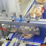 Zbliżenie na Automat pakujący poziomy FLOWPACK SBM550S-LR zgrzewający plastikowe torebki ze stosami okrągłych płaskich chlebów lub chlebów pita na niebieskim przenośniku taśmowym w zakładzie przetwórstwa spożywczego.