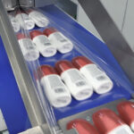 Automatyczna pakowarka flow-pack SR750LR owija kilka białych butelek z czerwonymi nakrętkami w przezroczysty plastik na przenośniku taśmowym w ramach zautomatyzowanego procesu pakowania w fabryce.