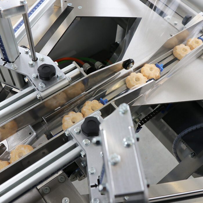Zbliżenie na zautomatyzowaną maszynę i Automatyczną pakowarkę flow-pack SR550E układającą ciastka w kształcie łapek na linii produkcyjnej, z metalowymi prowadnicami i przezroczystymi panelami kierującymi ciastka przez cały proces.